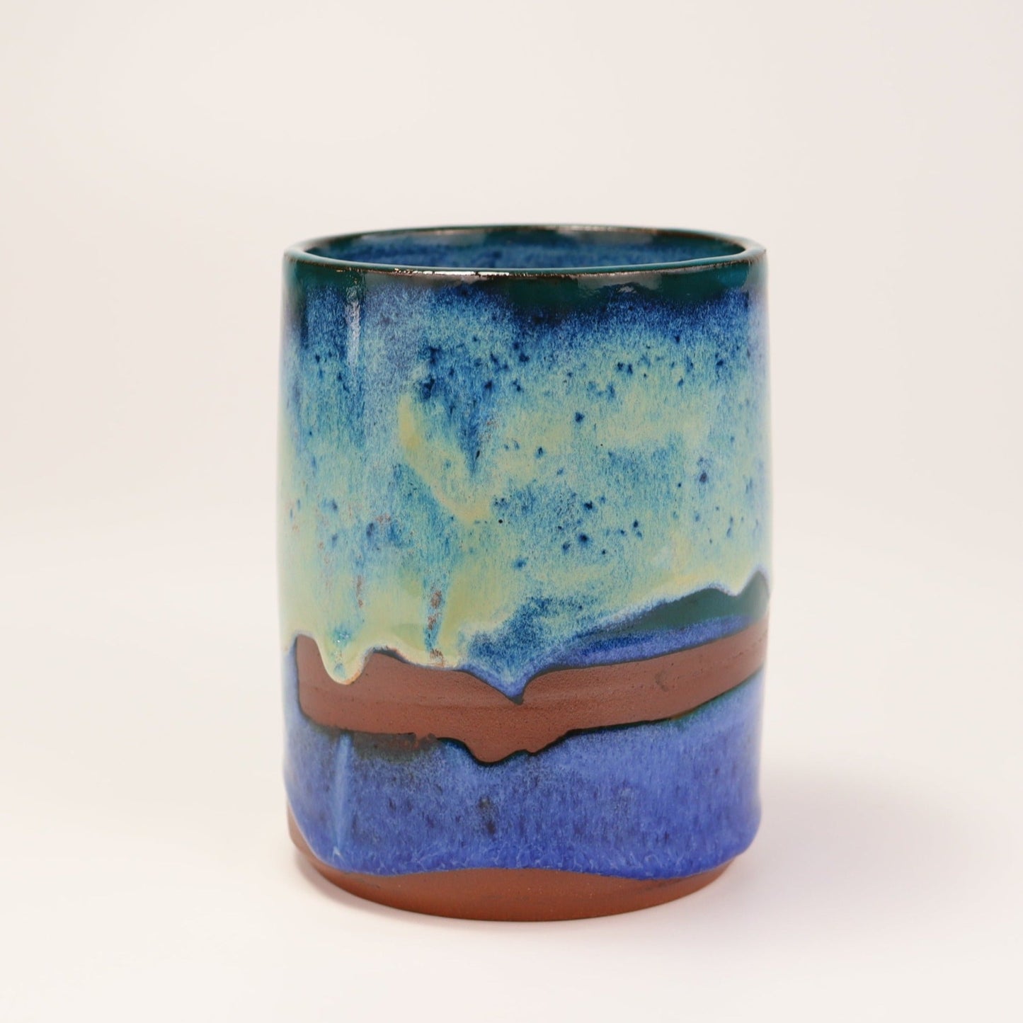 Handmade, wheel thrown highball style cup glazed in blues, teals and greens. I've deliberately left bare bands in a random fashion around the middle of the pot to reveal he deep red/burgundy clay body in a unique bikini-esque style.