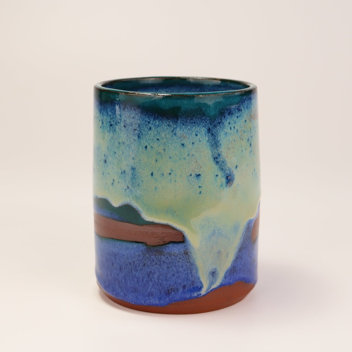 Handmade, wheel thrown highball style cup glazed in blues, teals and greens. I've deliberately left bare bands in a random fashion around the middle of the pot to reveal he deep red/burgundy clay body in a unique bikini-eques style.