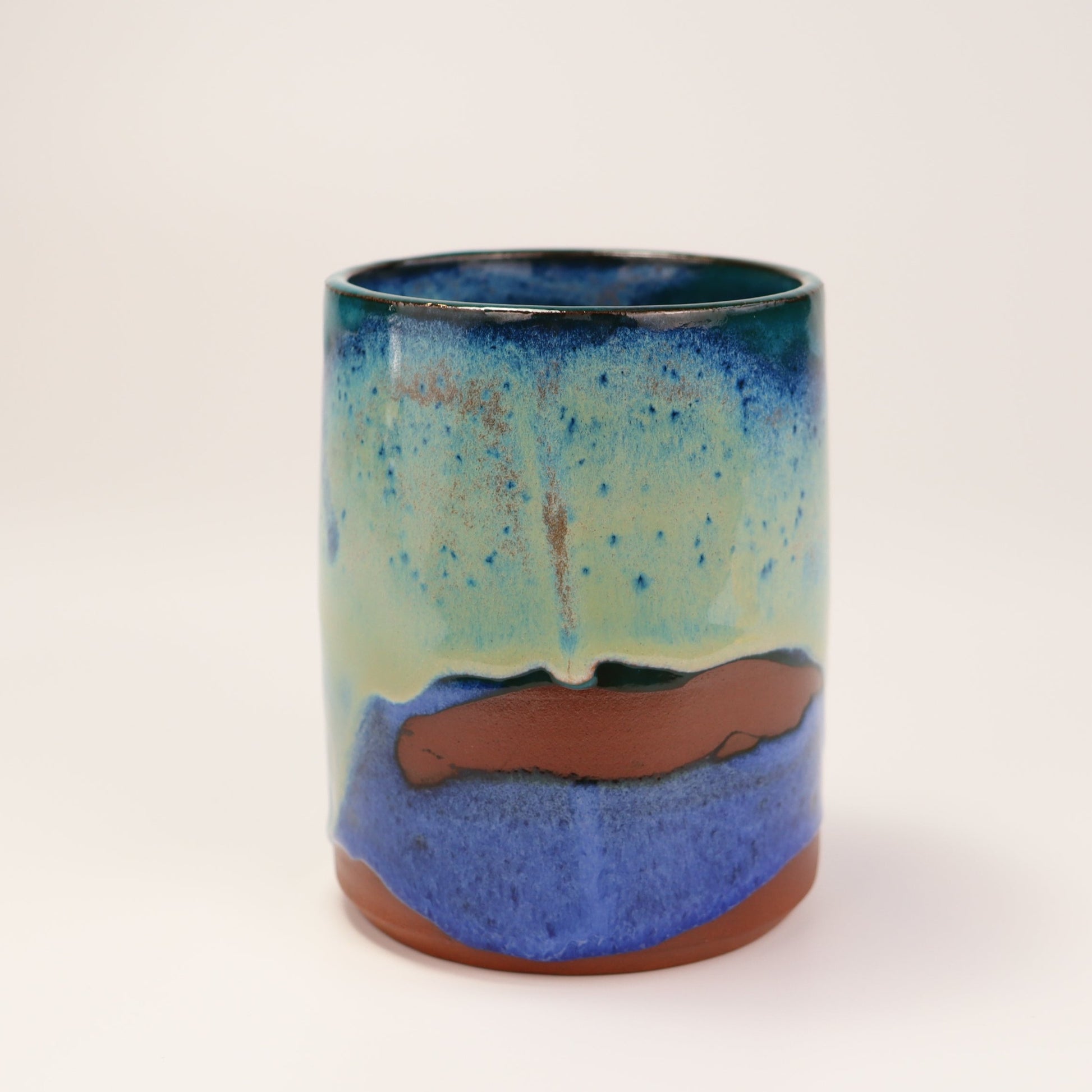 Handmade, wheel thrown highball style cup glazed in blues, teals and greens. I've deliberately left bare bands in a random fashion around the middle of the pot to reveal he deep red/burgundy clay body in a unique bikini-eques style.