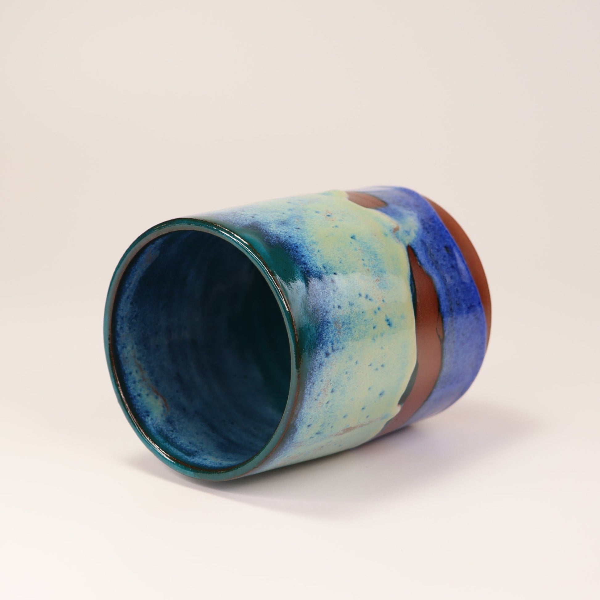 Handmade, wheel thrown highball style cup glazed in blues, teals and greens. I've deliberately left bare bands in a random fashion around the middle of the pot to reveal he deep red/burgundy clay body in a unique bikini-eques style.