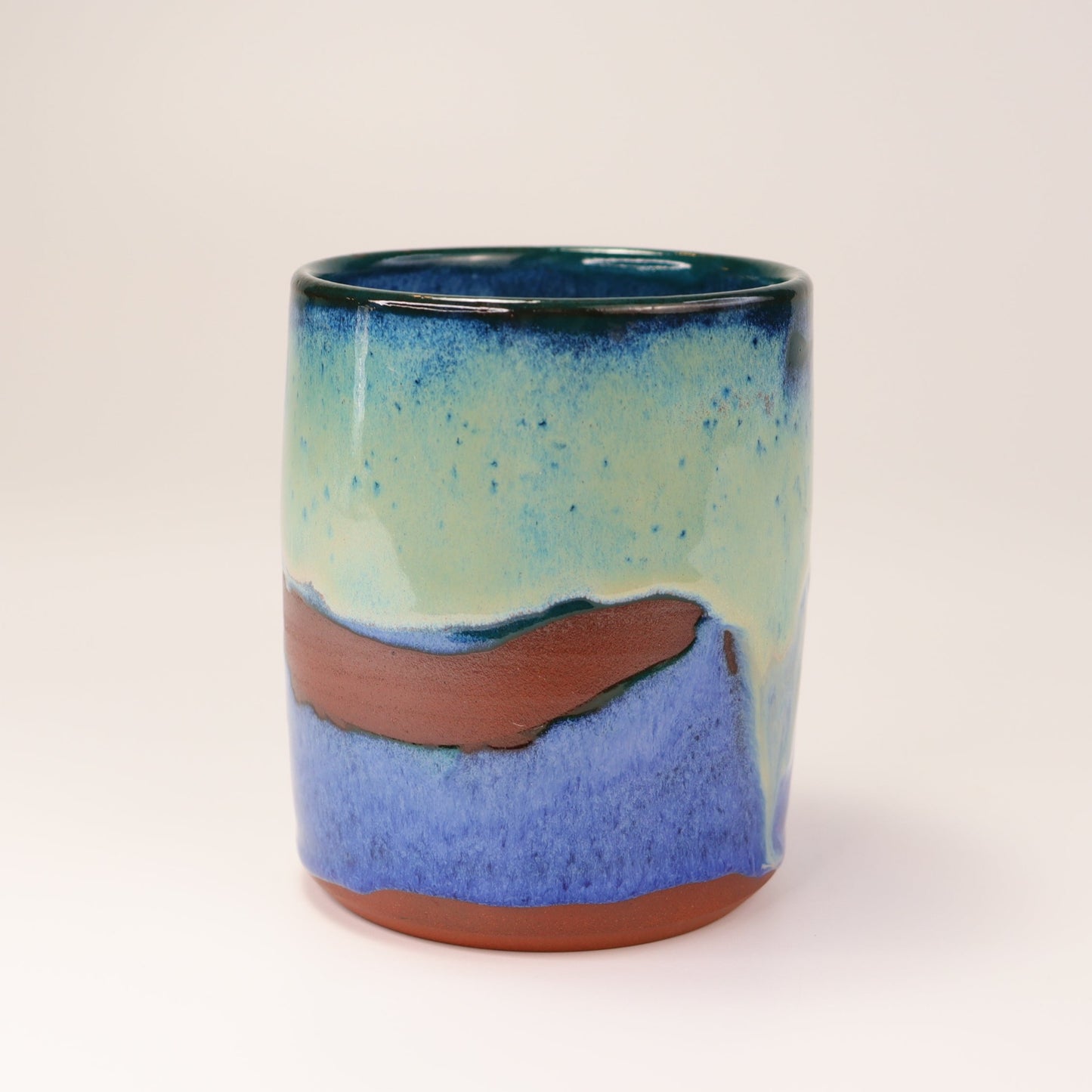 Handmade, wheel thrown highball style cup glazed in blues, teals and greens. I've deliberately left bare bands in a random fashion around the middle of the pot to reveal he deep red/burgundy clay body in a unique bikini-eques style.