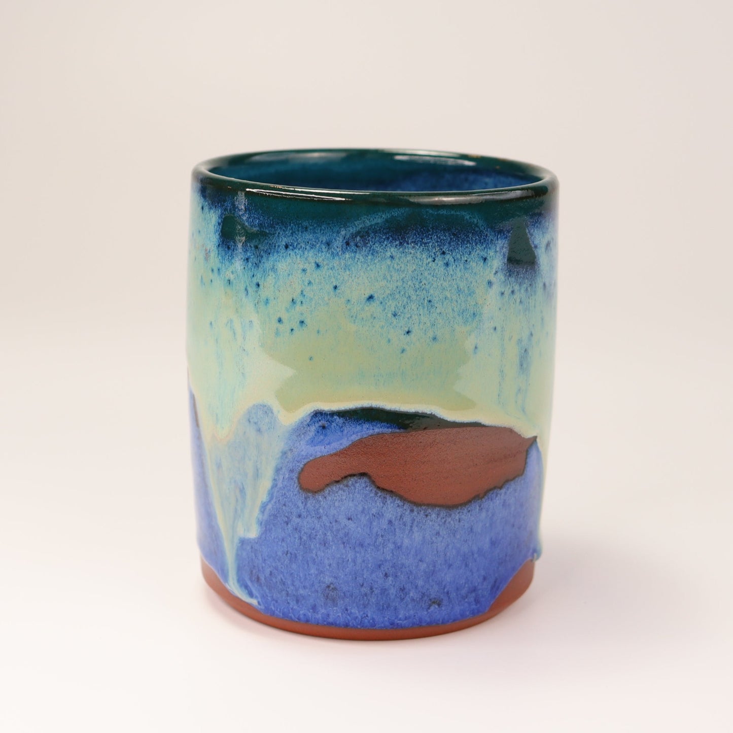 Handmade, wheel thrown highball style cup glazed in blues, teals and greens. I've deliberately left bare bands in a random fashion around the middle of the pot to reveal he deep red/burgundy clay body in a unique bikini-eques style.