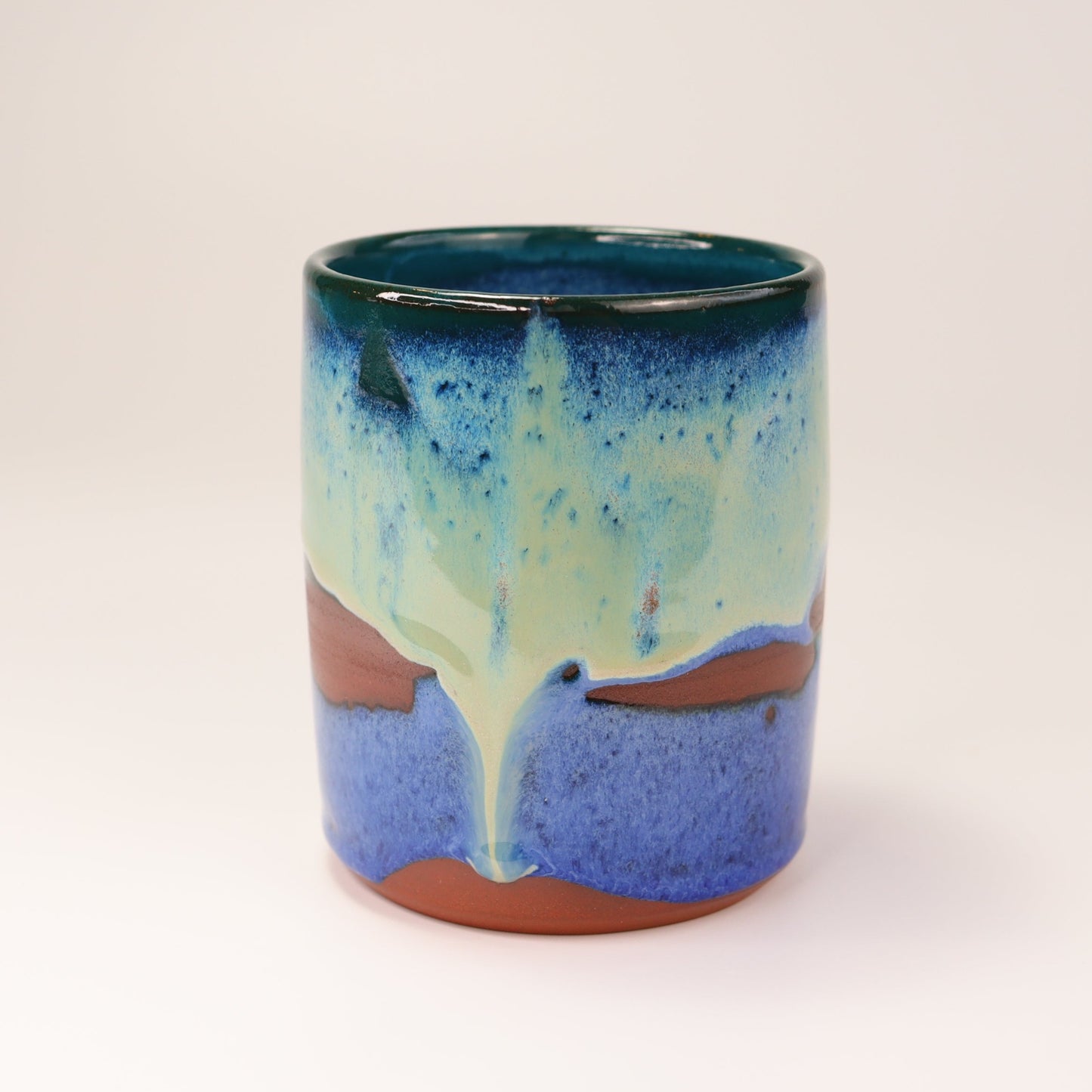 Handmade, wheel thrown highball style cup glazed in blues, teals and greens. I've deliberately left bare bands in a random fashion around the middle of the pot to reveal he deep red/burgundy clay body in a unique bikini-eques style.