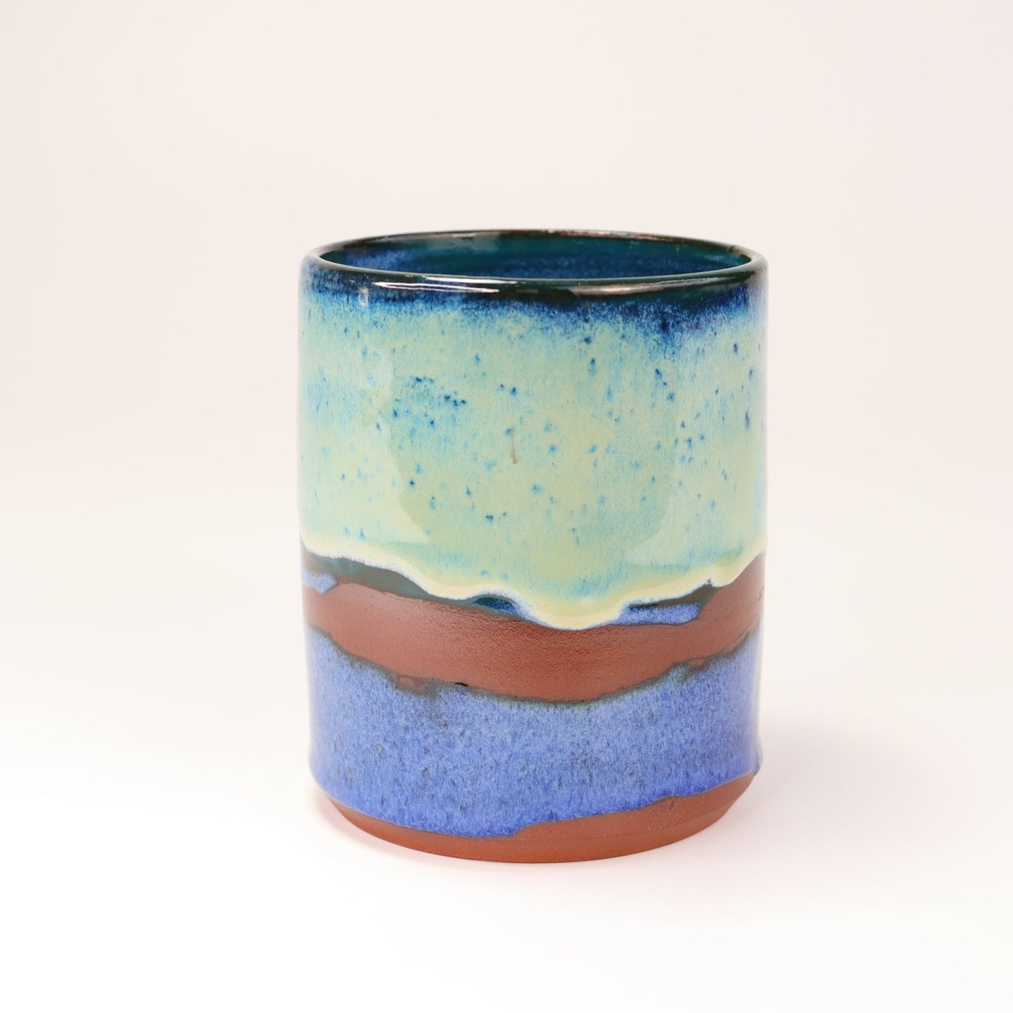 Handmade, wheel thrown highball style cup glazed in blues, teals and greens. I've deliberately left bare bands in a random fashion around the middle of the pot to reveal he deep red/burgundy clay body in a unique bikini-eques style.